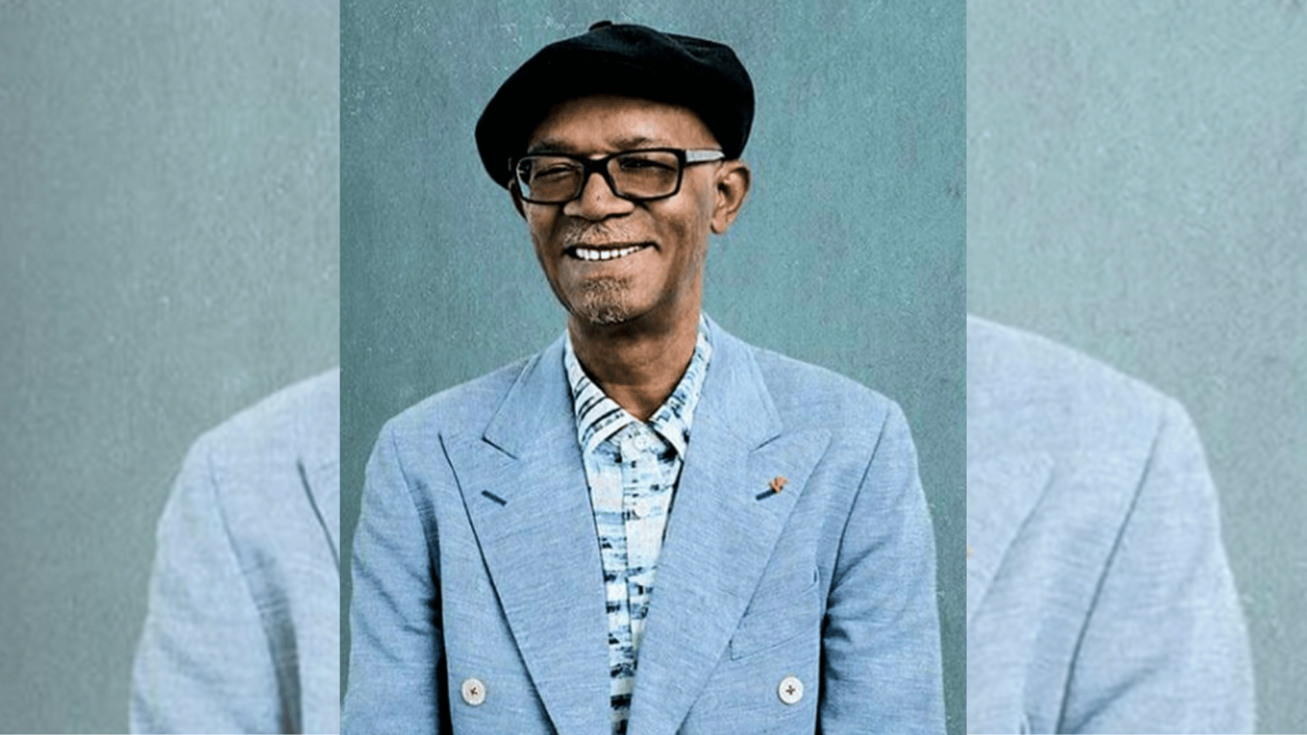 smiling man in glasses, beret, and pale blue suit posed against textured blue backdrop.