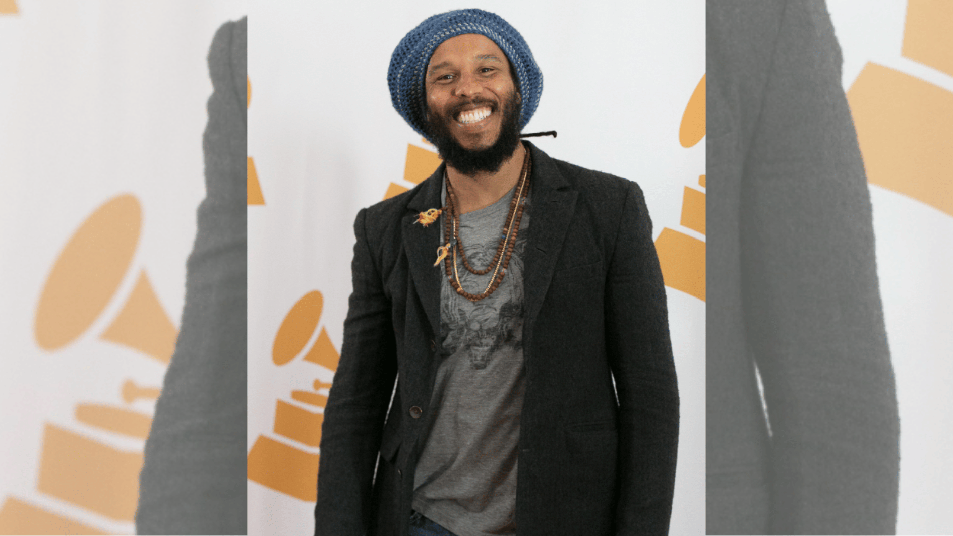 smiling reggae artist in blue knit tam and black blazer posing at awards event backdrop.