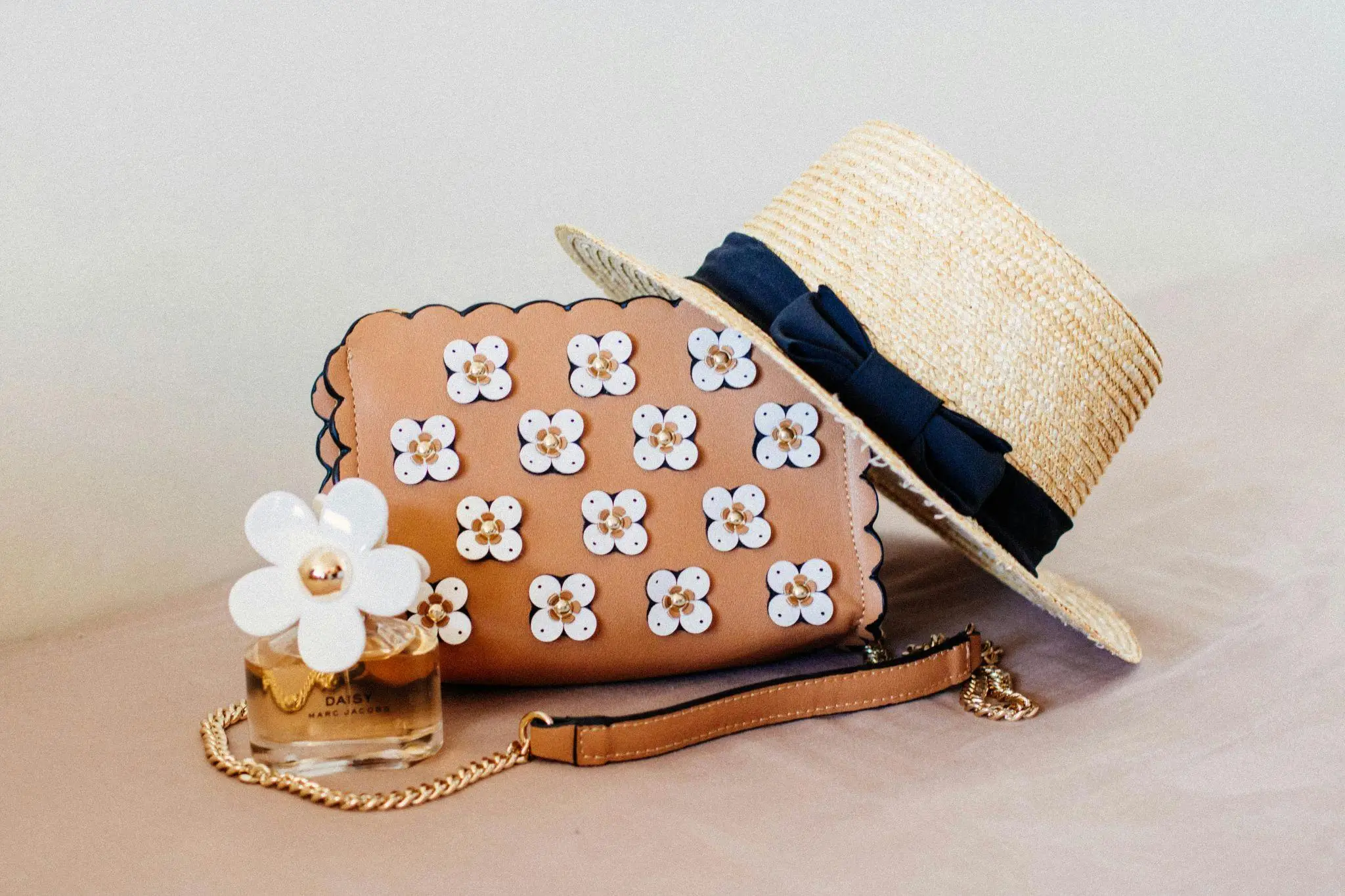 Beige handbag with floral embellishments, straw hat, and perfume bottle on soft surface