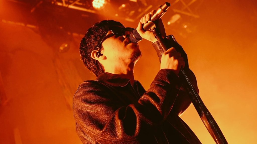 young male singer performing on stage with a microphone under bright orange stage lighting wearing a leather jacket