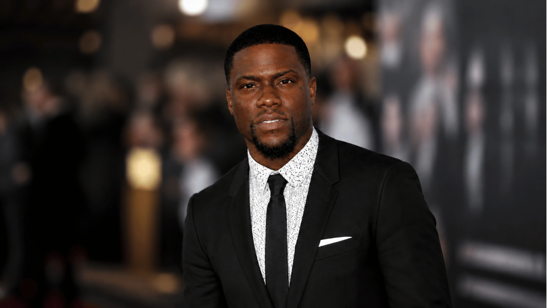 Professional portrait of Kevin Hart in a black suit at a movie premiere event with red carpet lights and crowd in the background.