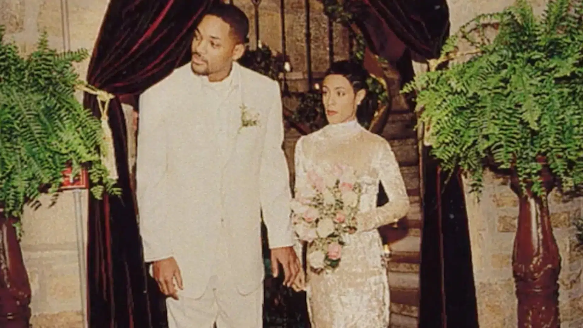 Will Smith and Jada Pinkett Smith on their wedding day, groom in white suit, bride in lace gown holding bouquet.