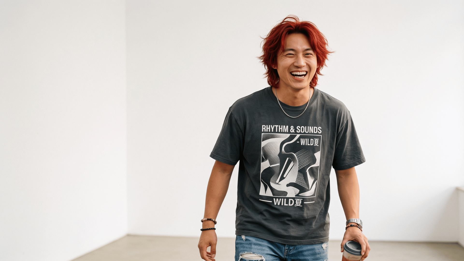 a laughing man with red hair wears a dark graphic t shirt and jeans while holding a cup in a plain white room