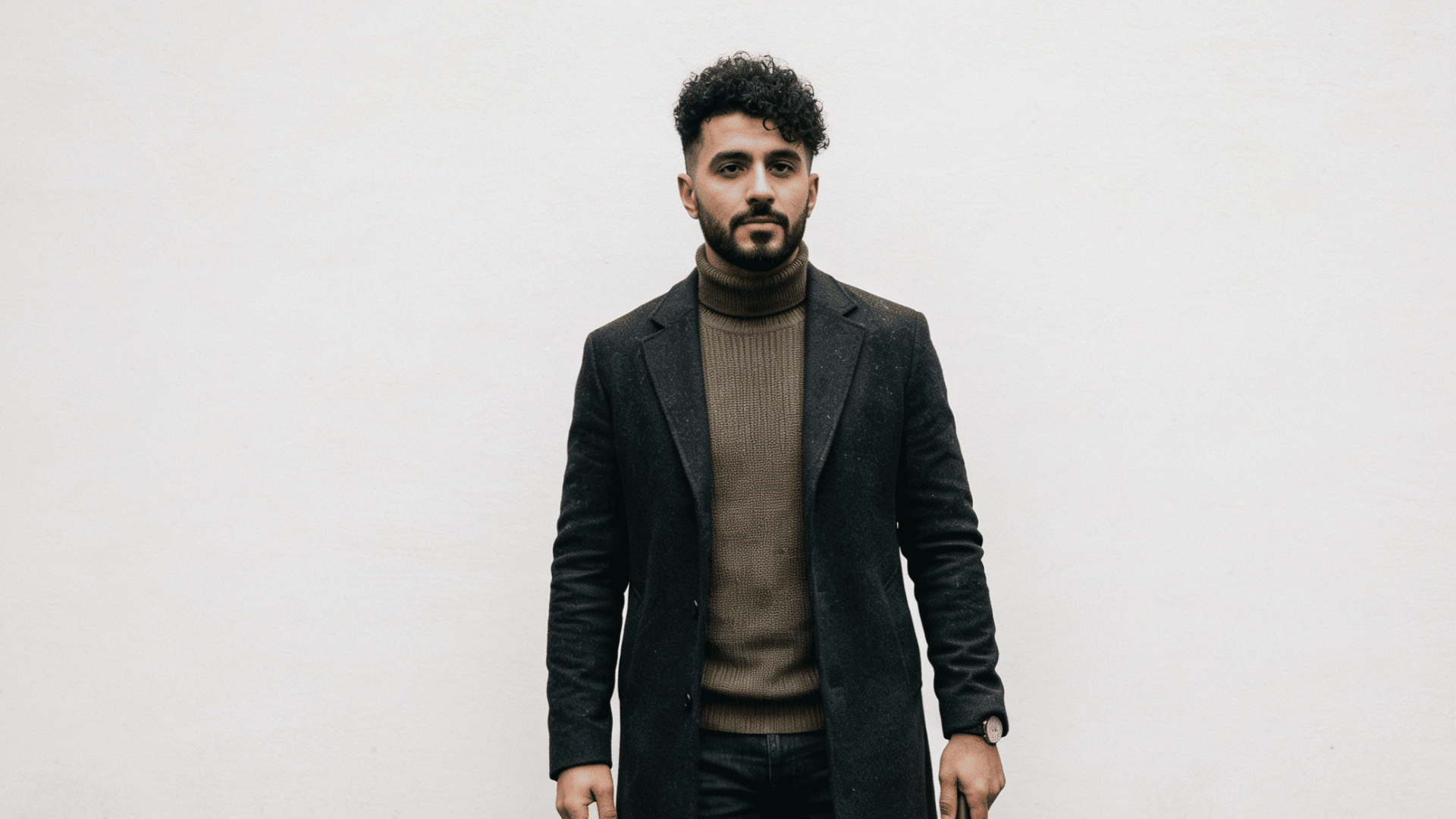 a man with curly hair wearing a dark coat and olive turtleneck stands against a plain white wall holding an umbrella