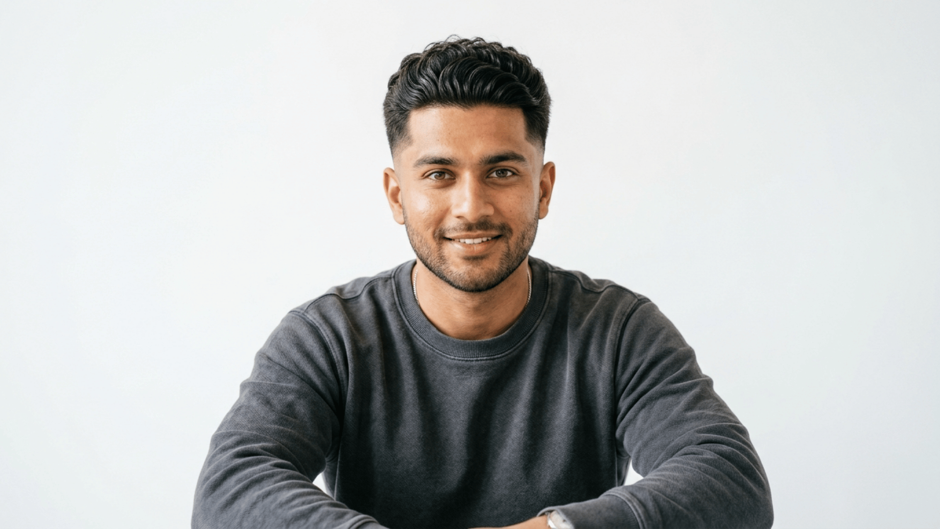 a smiling man with styled dark hair wears a gray sweatshirt while leaning forward against a plain light background