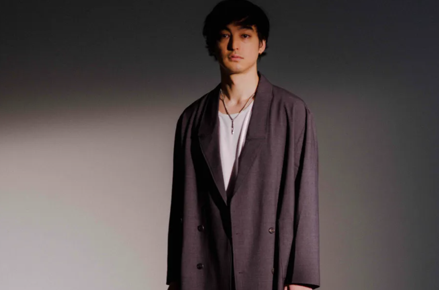 a young man in a dark oversized blazer over a white shirt standing against a plain gray studio background