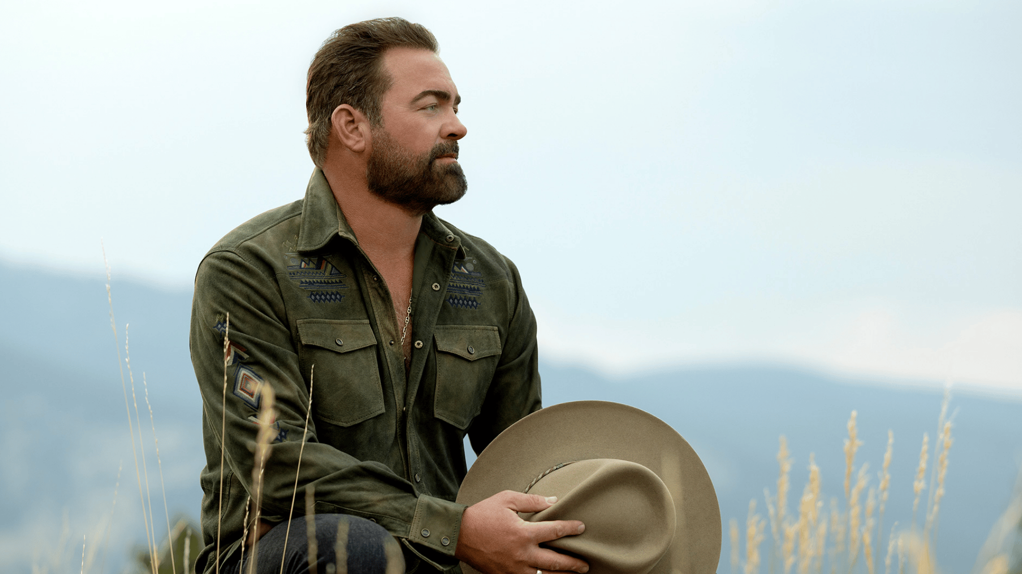 country singer sitting outdoors holding a cowboy hat while wearing a green western-style jacket in nature