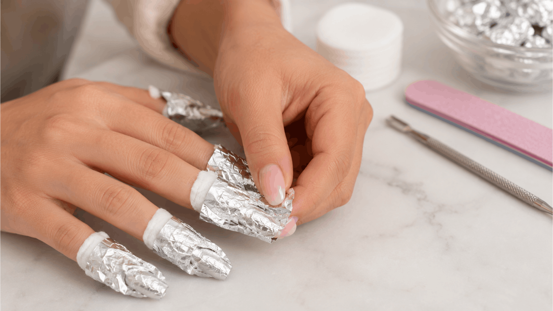 close up of hands with gel nails wrapped in foil and cotton pads for removal on a white marble surface with manicure tools nearby