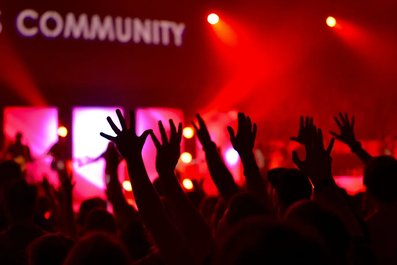 Concert audience with raised hands under red stage lighting in a crowded venue