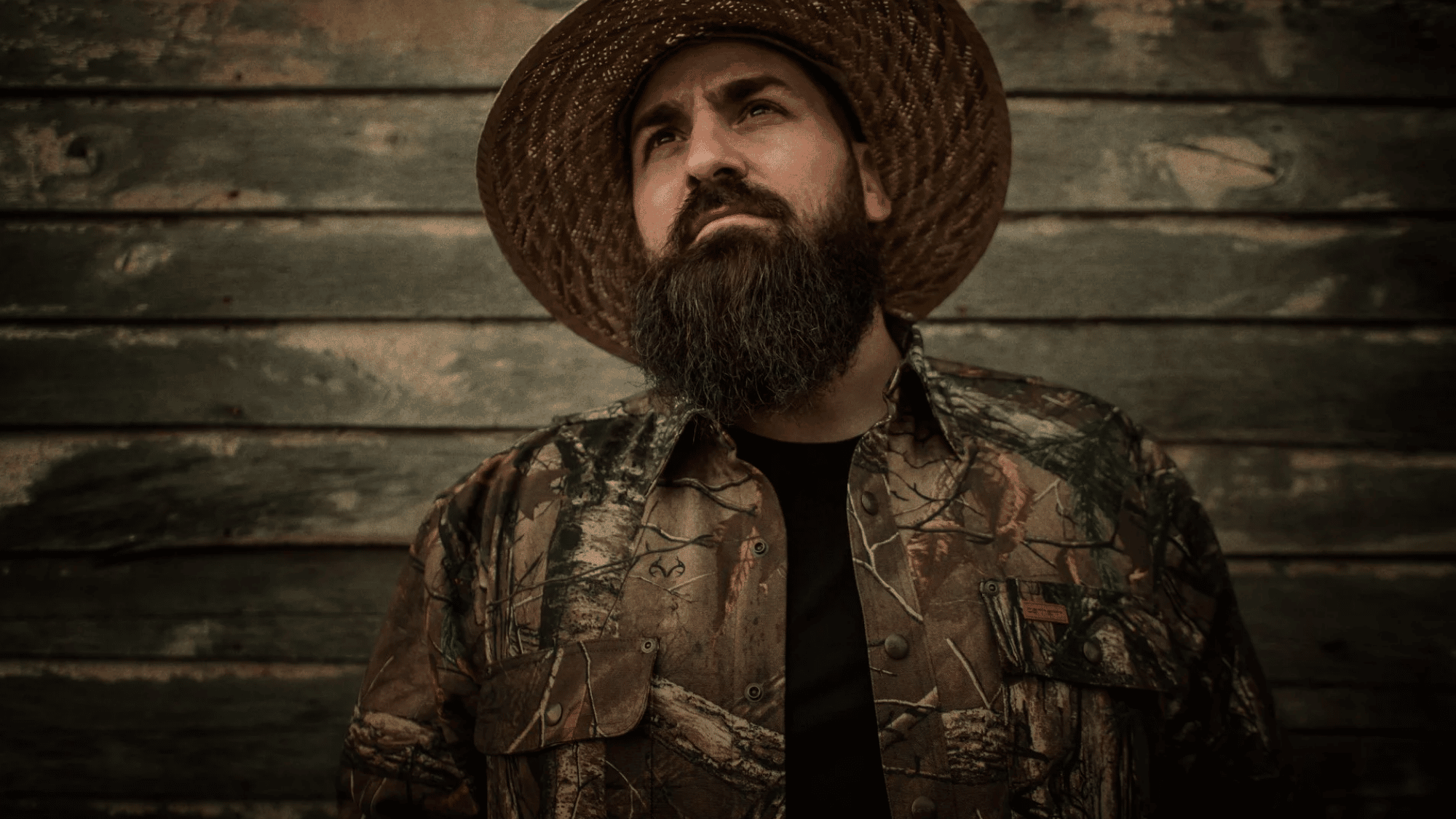 demun jones wearing a camouflage jacket and straw hat while posing against a rustic wooden backdrop