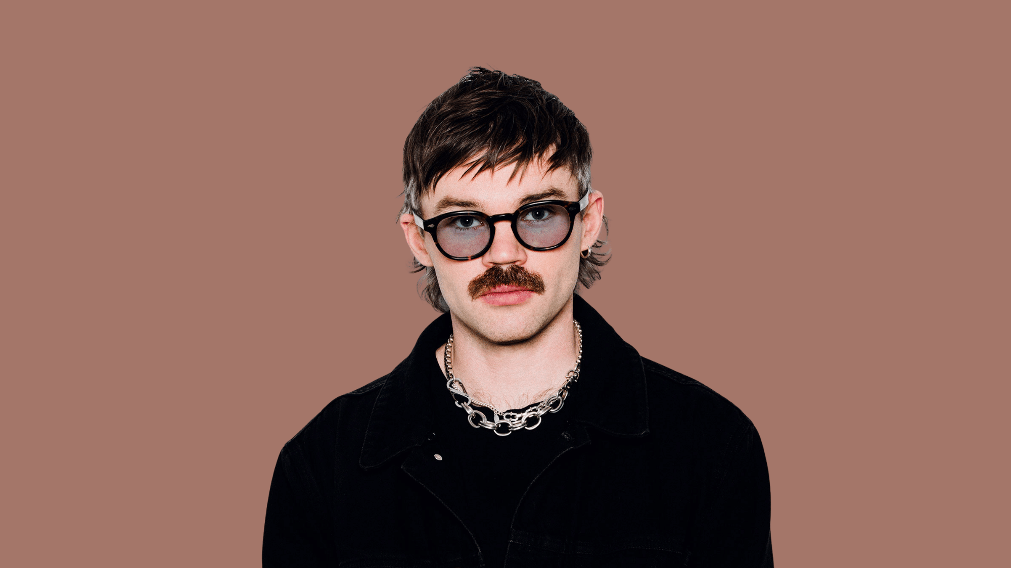Portrait of dom dolla with glasses, black jacket, and chain necklace on plain background.
