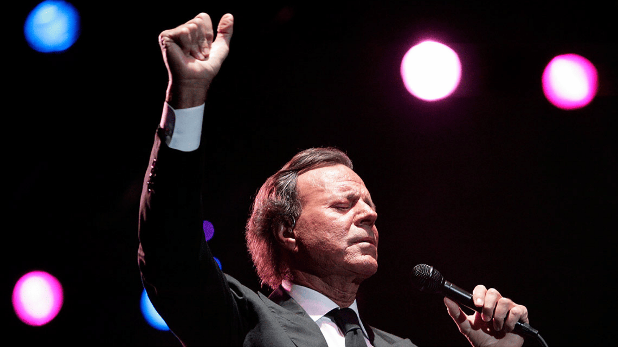 Julio Iglesias performing on stage, eyes closed, holding microphone with raised hand