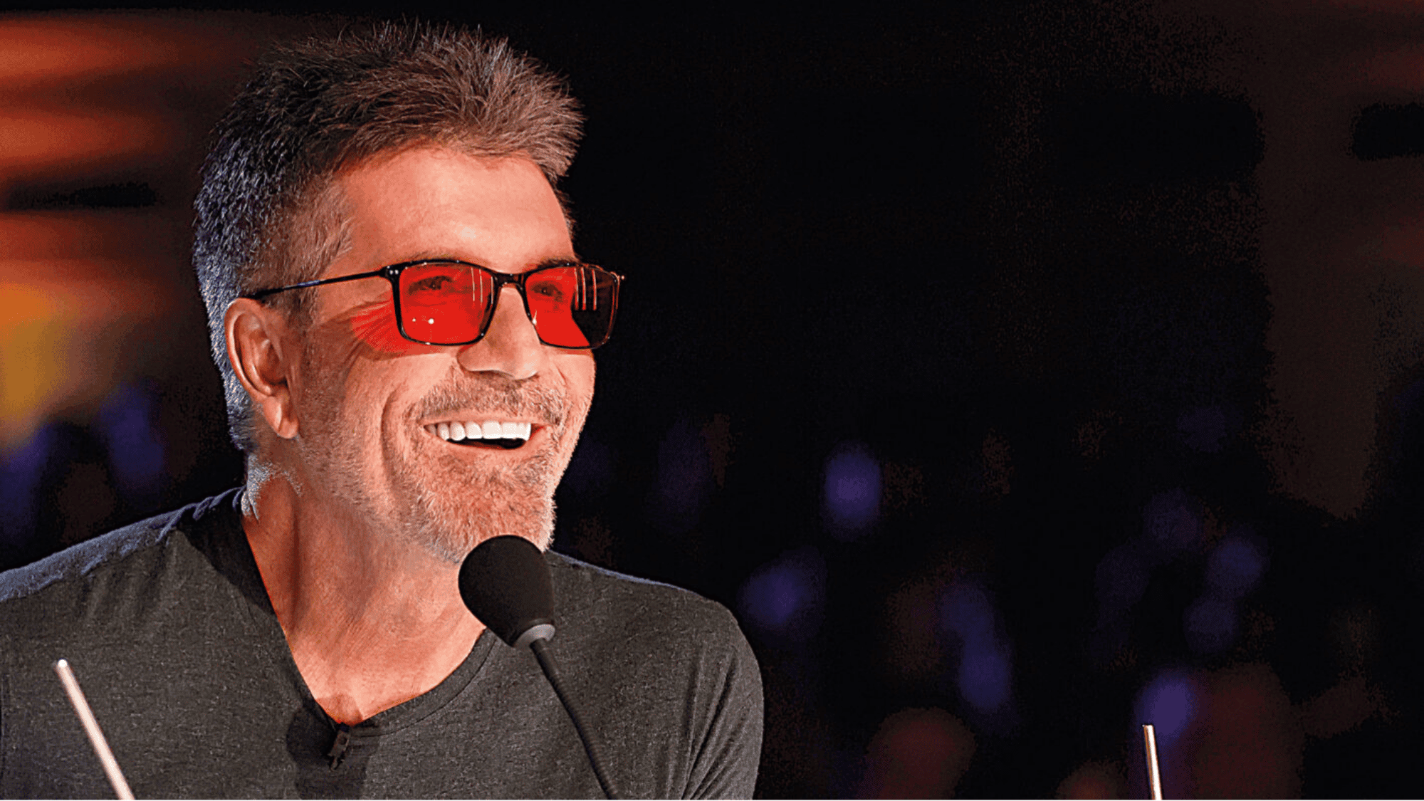 Simon Cowell smiling with red glasses and microphone at a live TV show