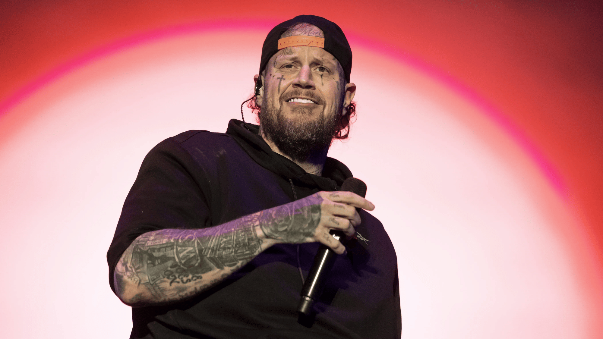 jelly roll performing live on stage holding a microphone while wearing a black hoodie and backward cap