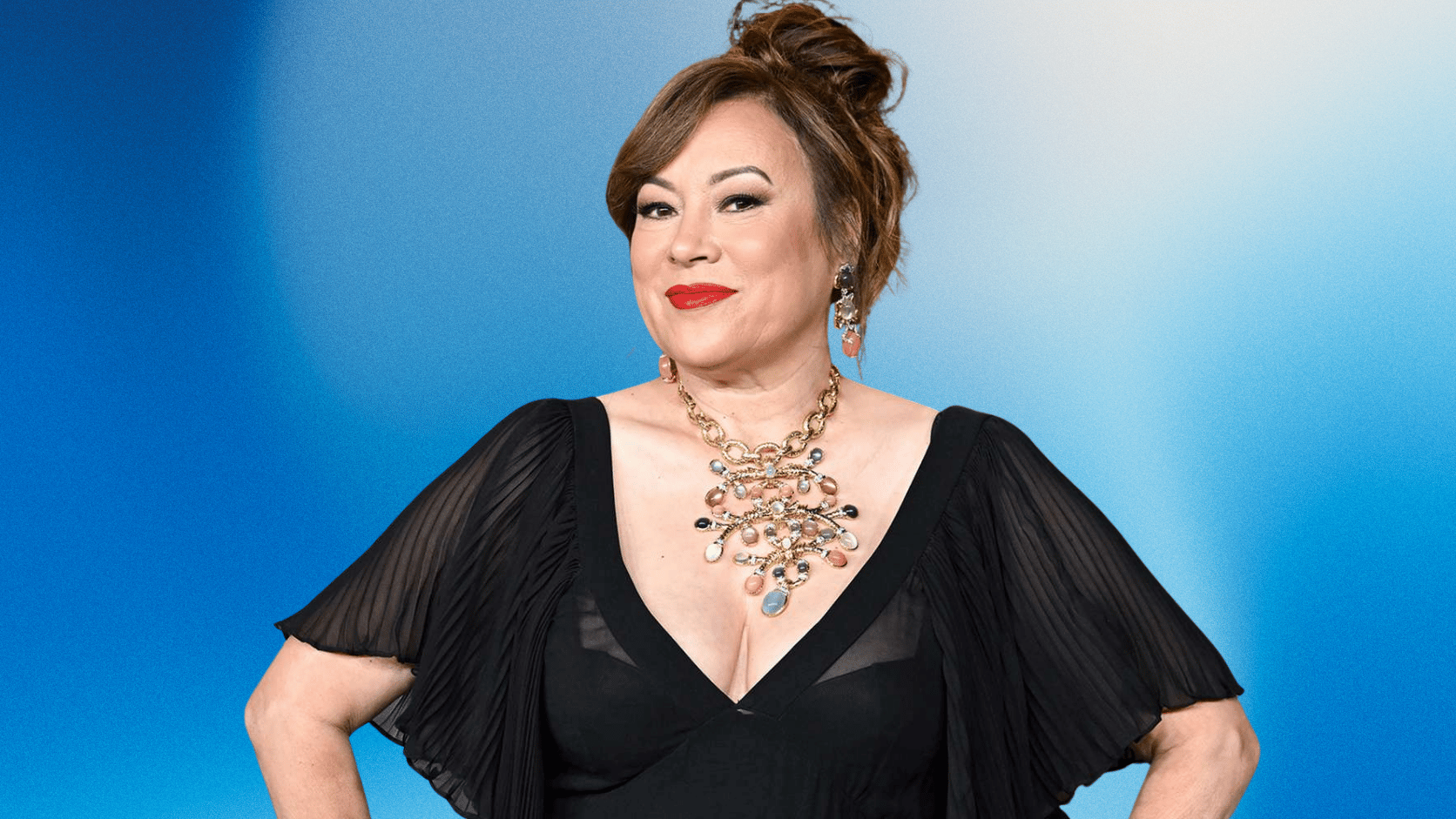Jennifer Tilly poses confidently in a black dress with statement jewelry against a blue gradient background.