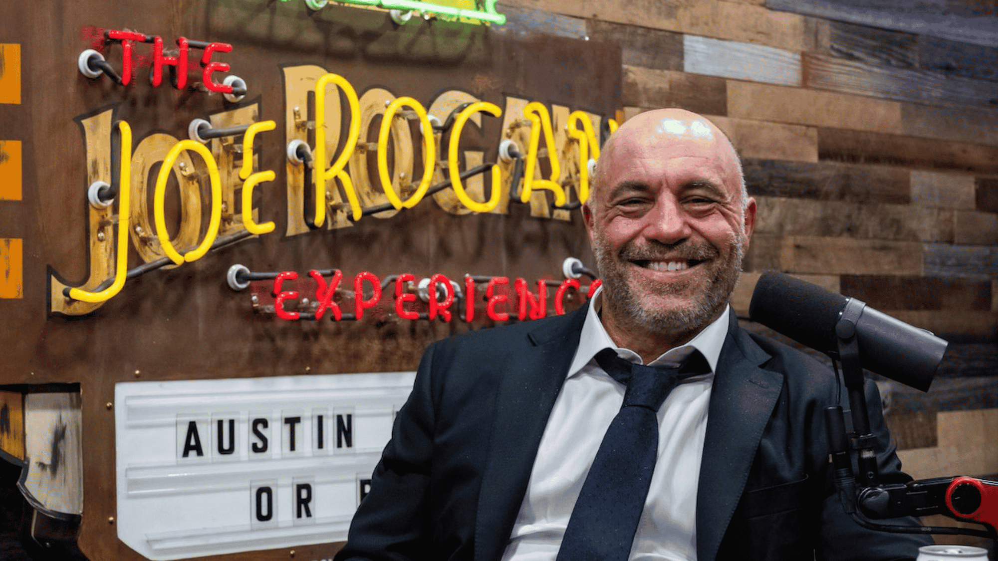 Joe Rogan laughing at his podcast studio in front of The Joe Rogan Experience sign