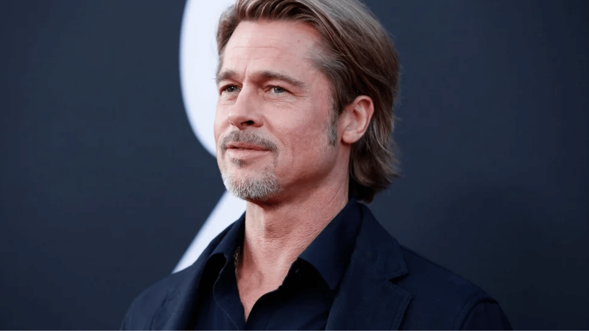 brad pitt posing at a red carpet event in a dark suit with styled hair and a blurred backdrop behind him