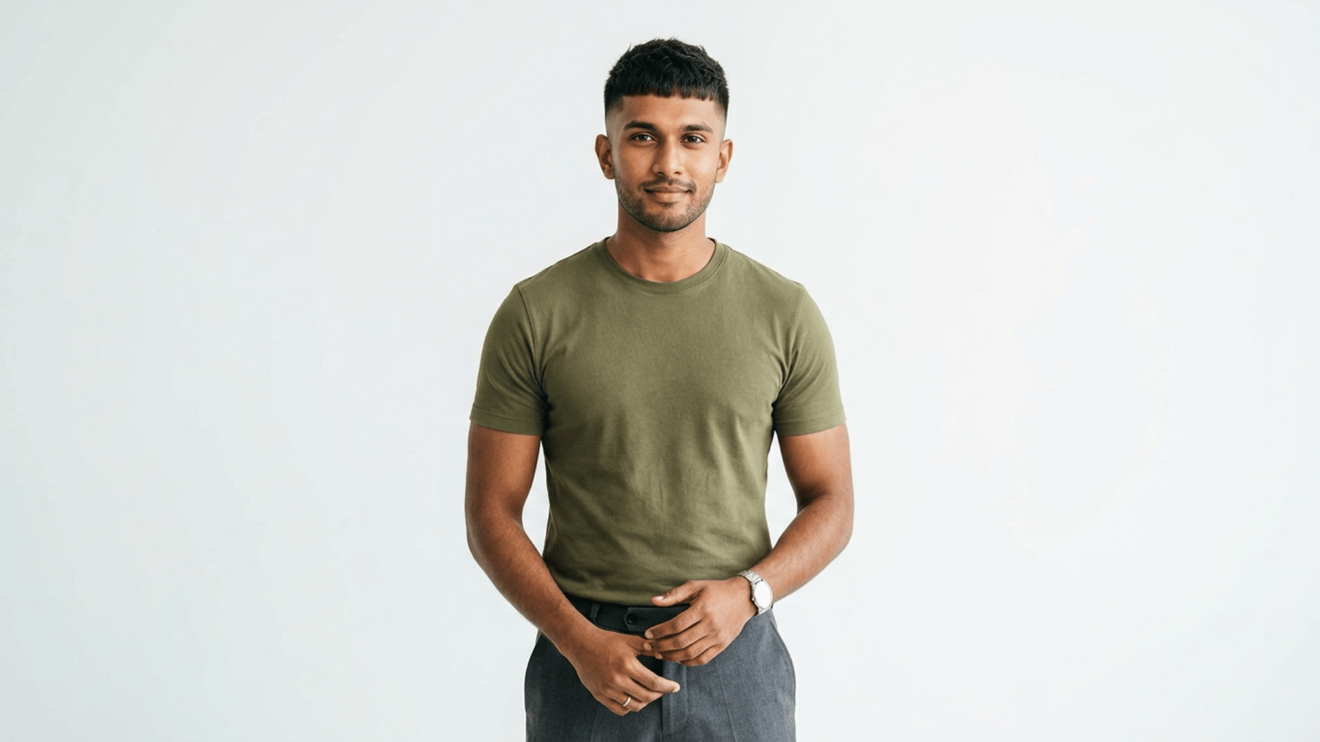 man standing against a plain light background wearing a green t shirt and gray trousers with hands gently clasped in front