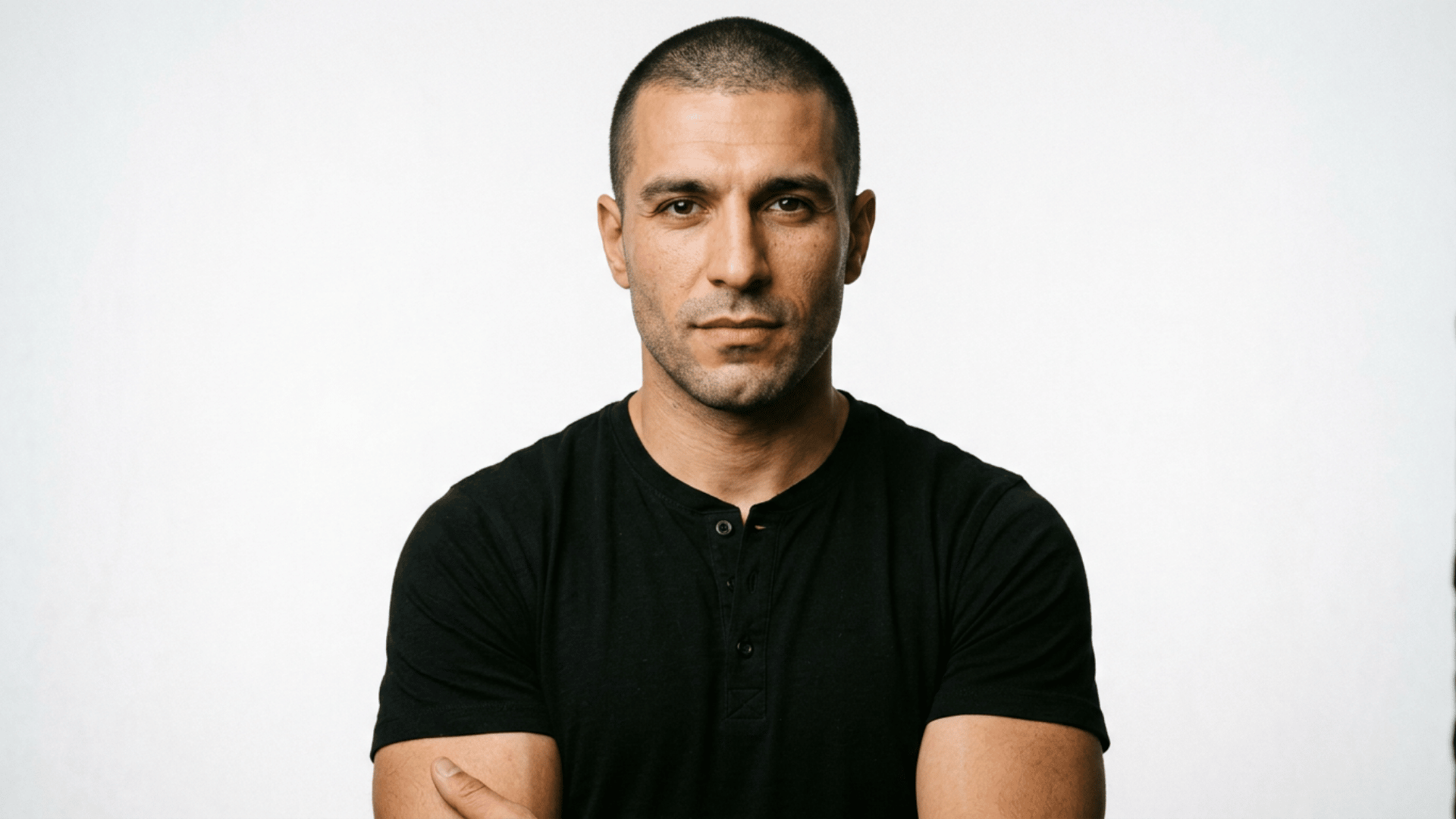 man with a shaved head wearing a black shirt posing with folded arms against a plain light background