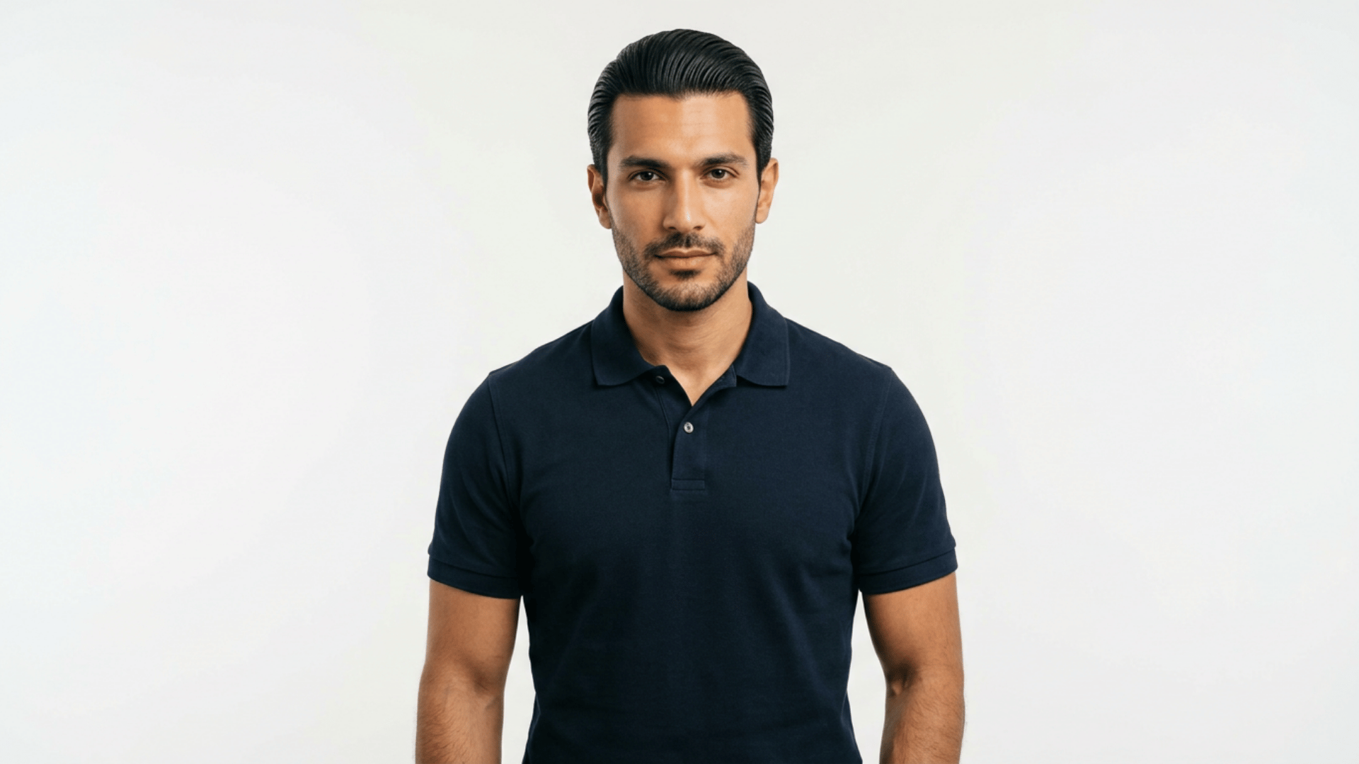 man with slicked back hairstyle and trimmed beard wearing a navy polo shirt standing confidently against a plain light background