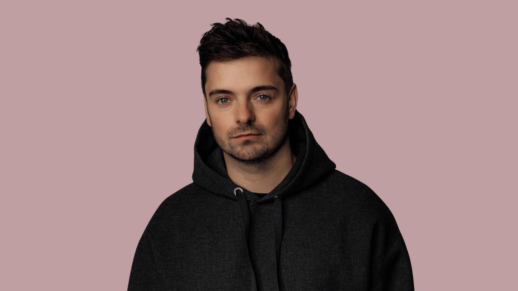 Portrait of martin garrix in dark hoodie with short styled hair against soft pink background.