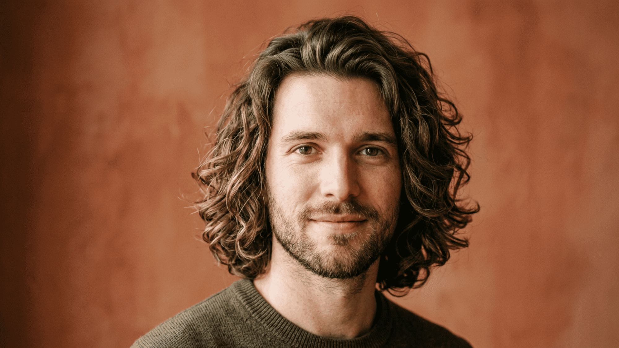 Man with oblong face styled with medium length wavy hair against warm background