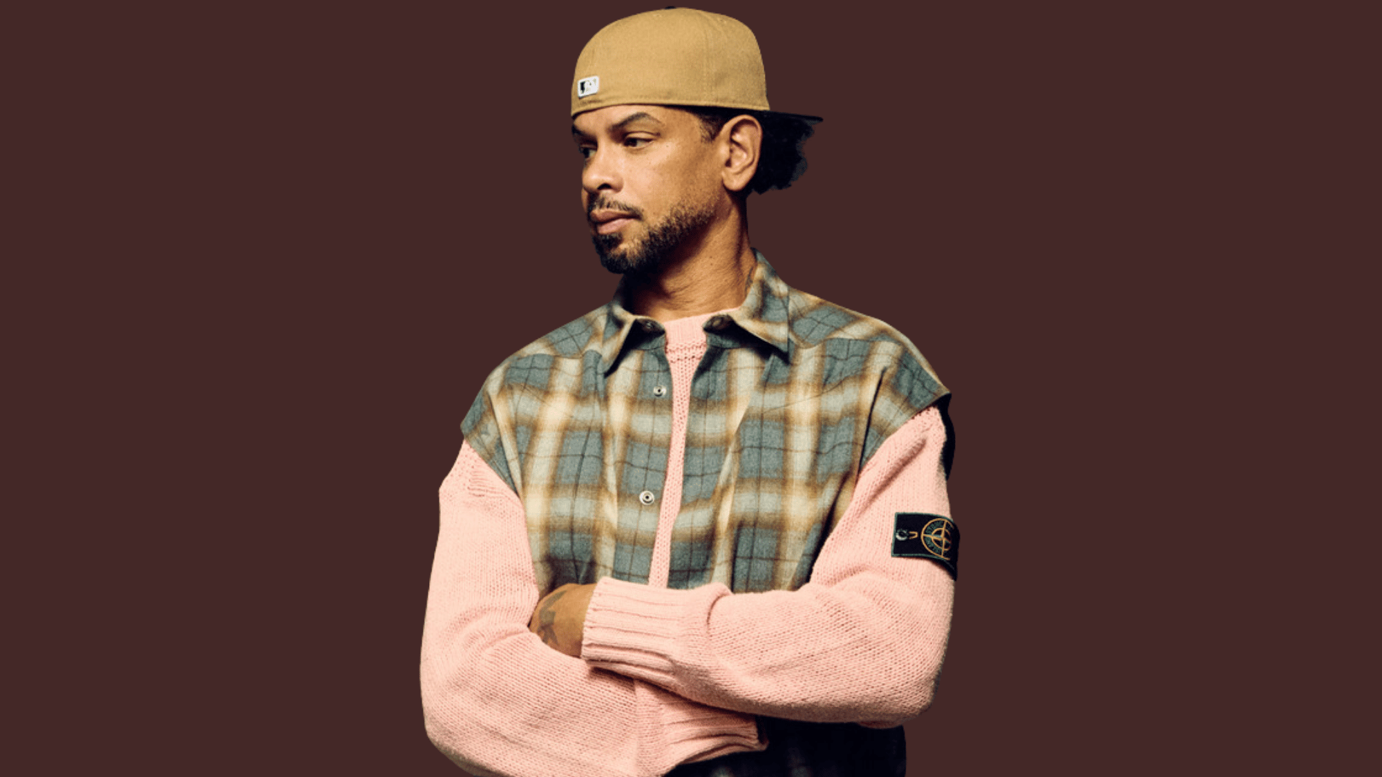 MK in beige cap wearing layered plaid vest over pink sweater, arms crossed, looking aside.