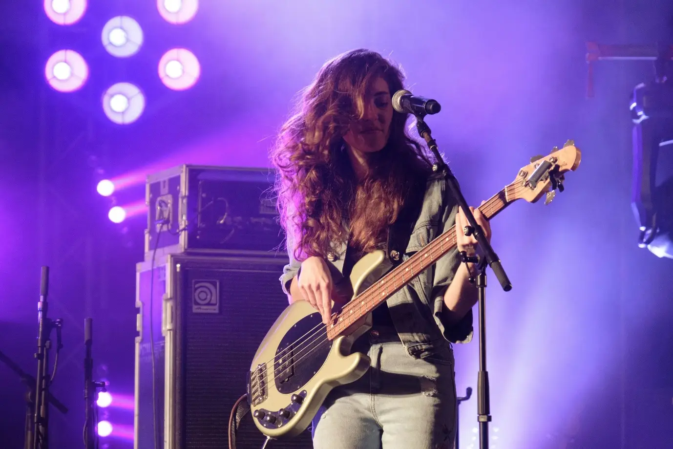 Musician playing bass guitar on stage under purple lighting with speakers in background