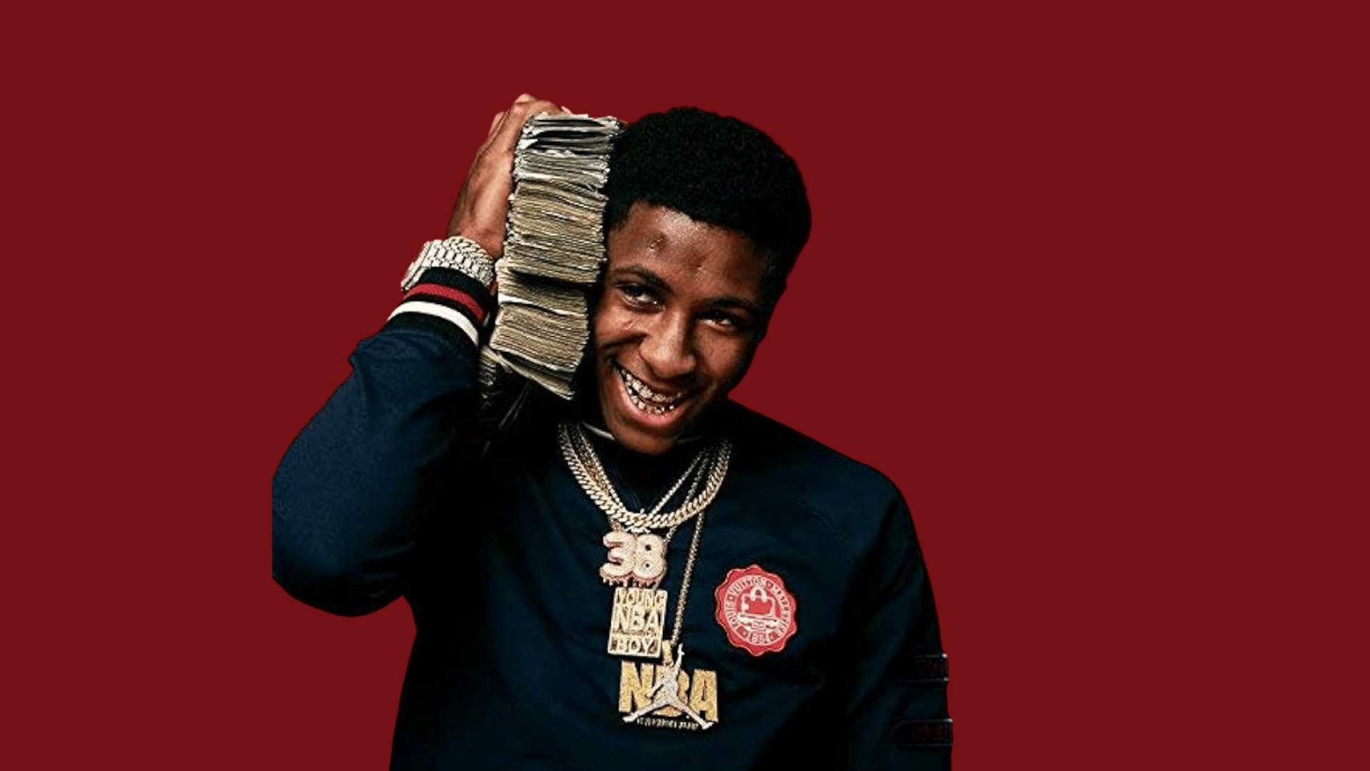 NBA YoungBoy holding a large stack of cash and wearing chains, smiling against a red background.