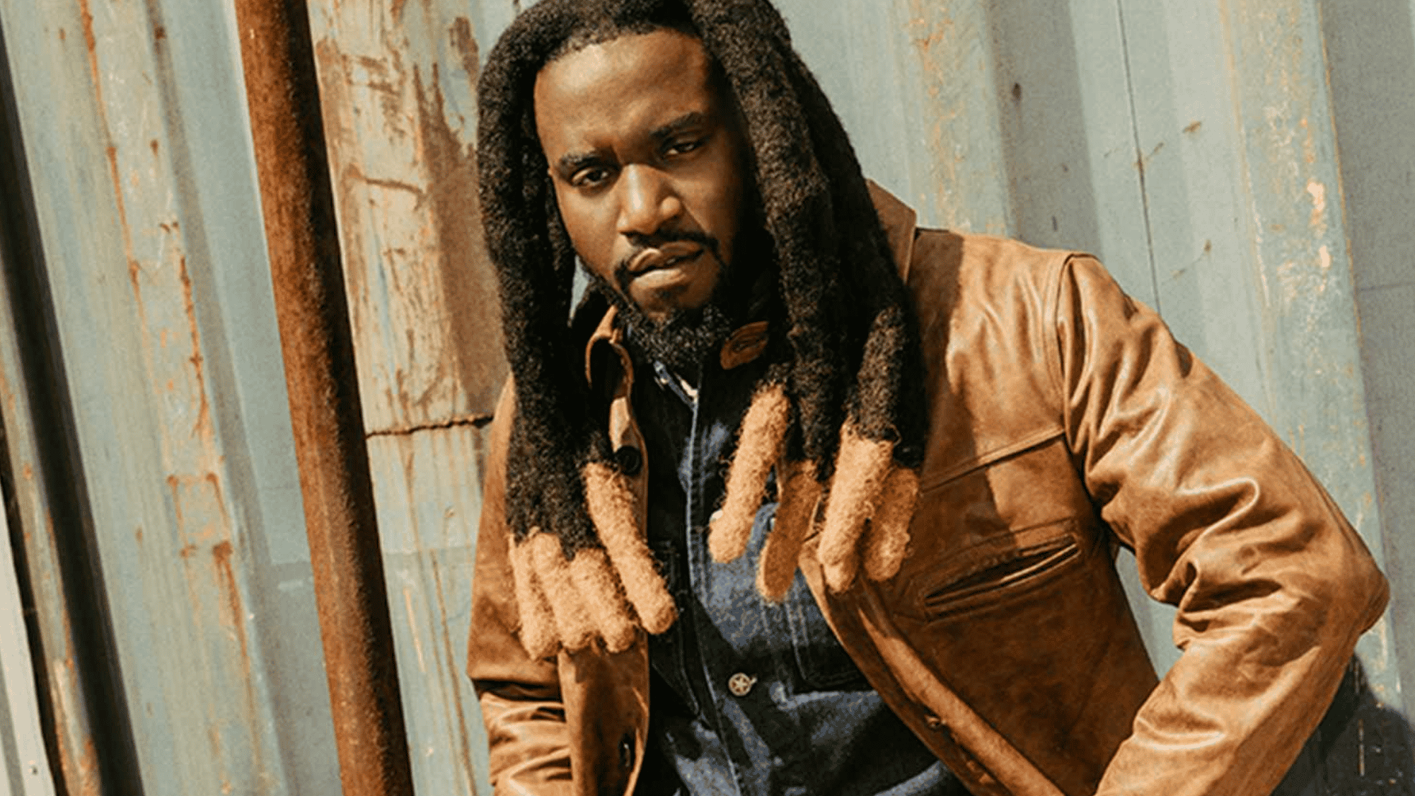 shaboozey posing outdoors in a brown leather jacket with long dreadlocks against a rustic metal backdrop