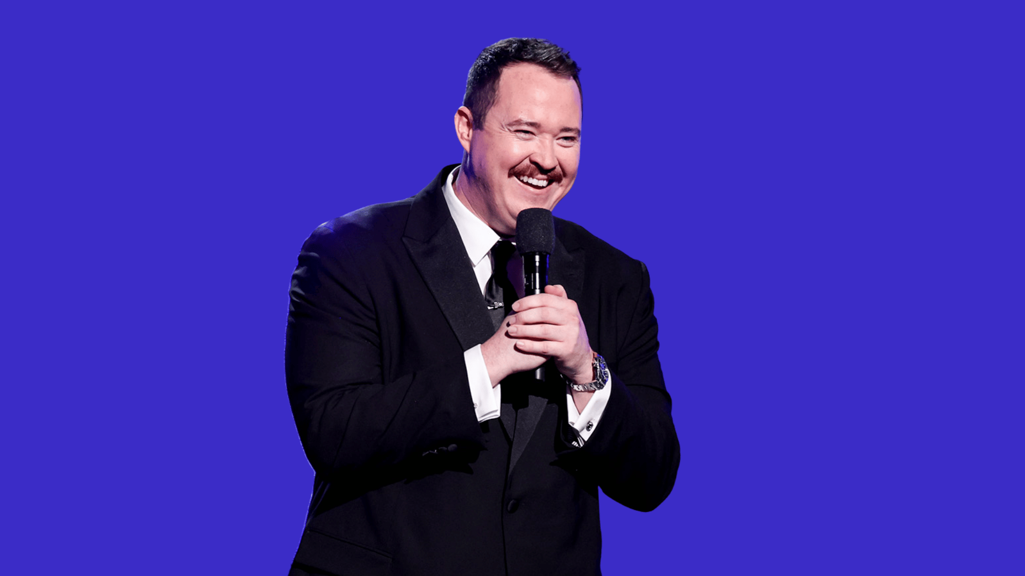 Shane Gillis standing with a mic against a purple background