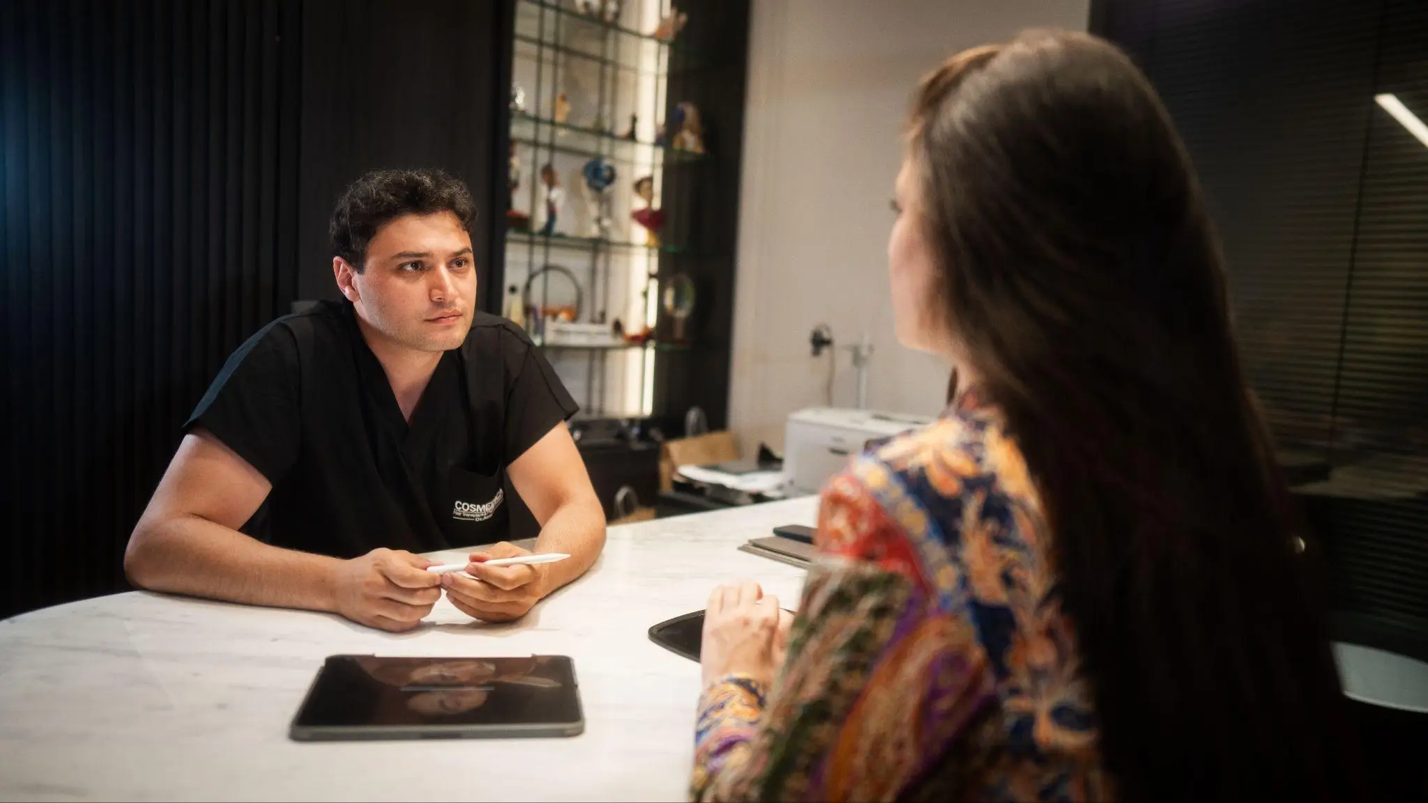 Doctor in black scrubs holding pen, discussing with woman in patterned blouse at white table