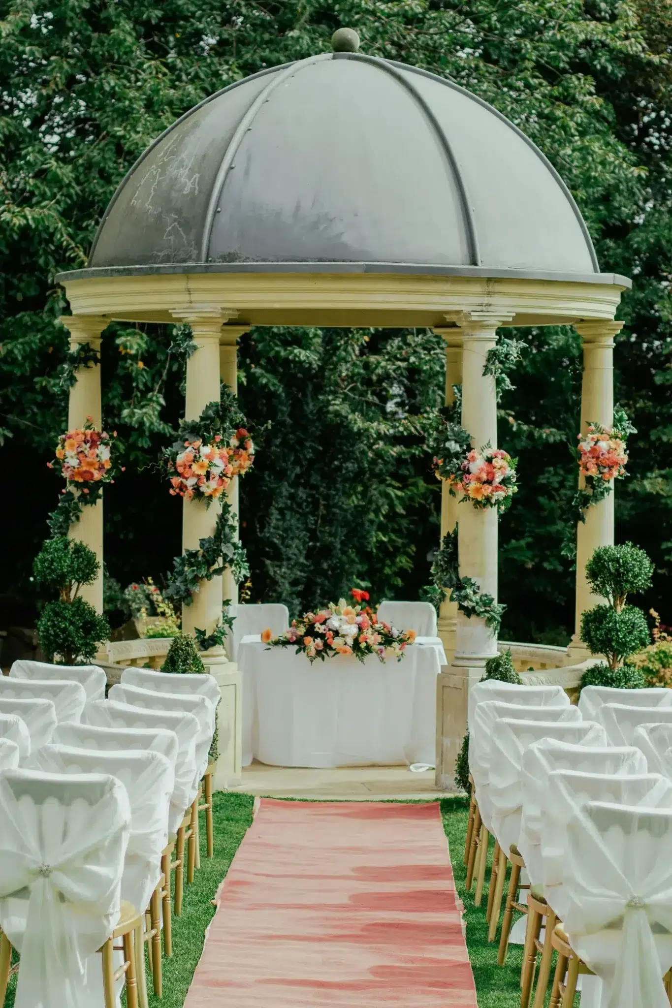 Outdoor wedding ceremony with white chairs and floral arch under dome in lush garden setting