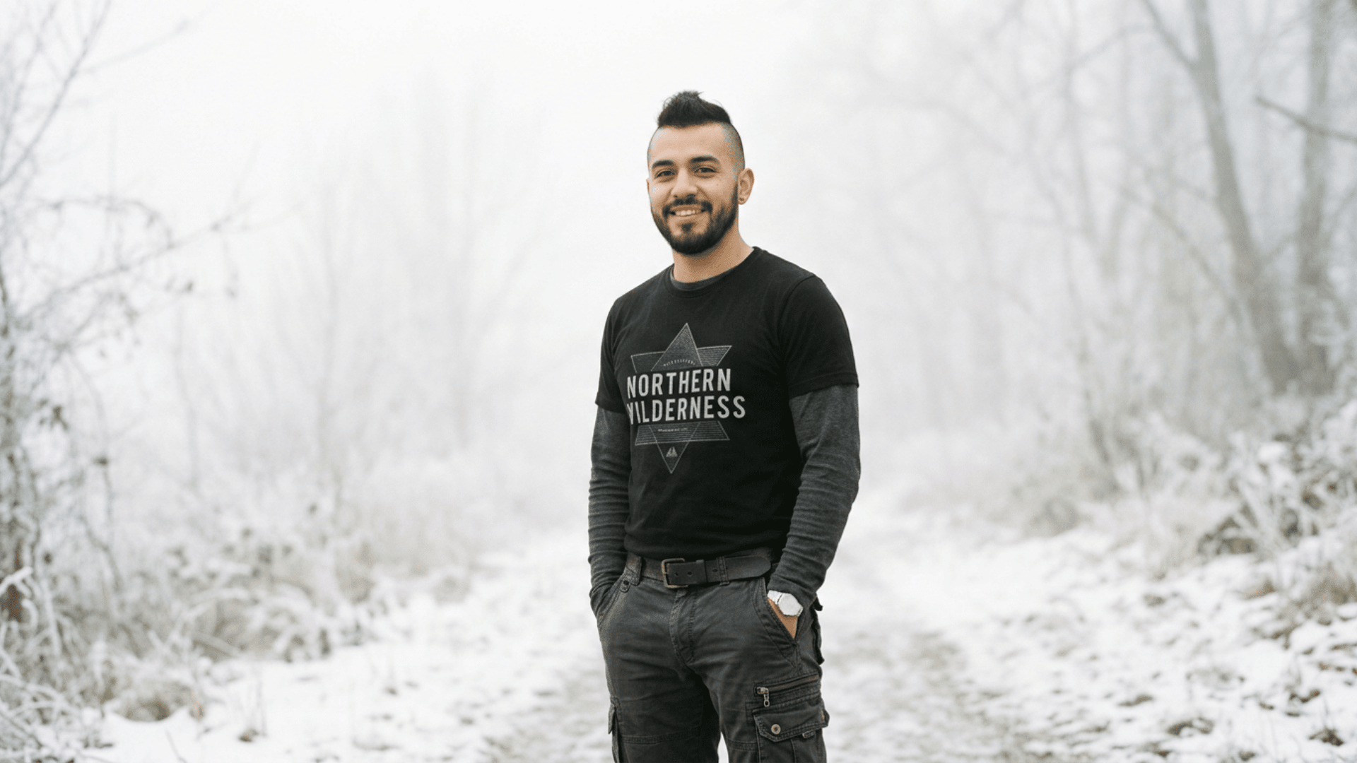 young man in a black graphic shirt standing on a snowy path surrounded by frost covered trees outdoors