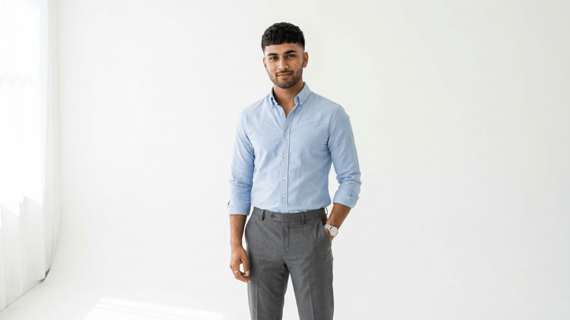 young man in a light blue shirt and gray trousers standing with one hand in his pocket against a bright background
