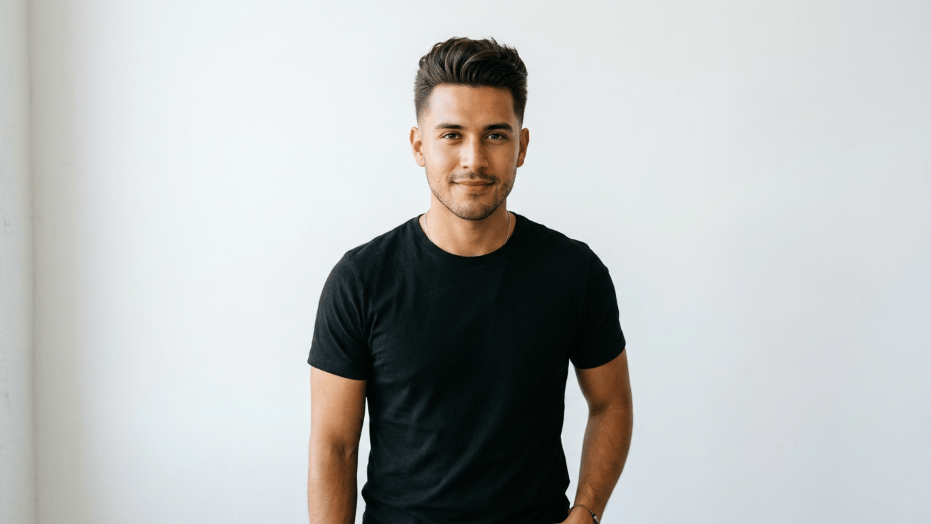 young man with a styled pompadour haircut wearing a black shirt standing confidently against a plain light wall background