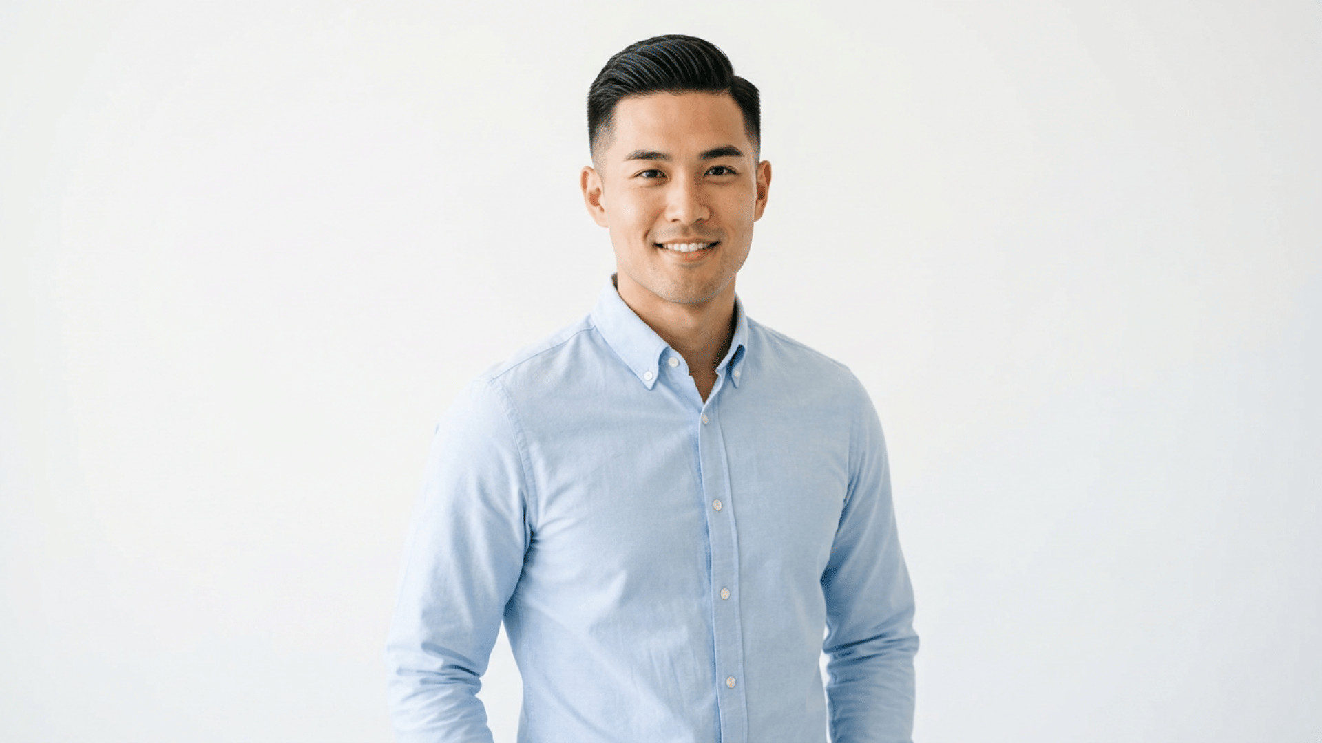 young man with neatly styled side parted haircut wearing a light blue shirt smiling against a plain light background