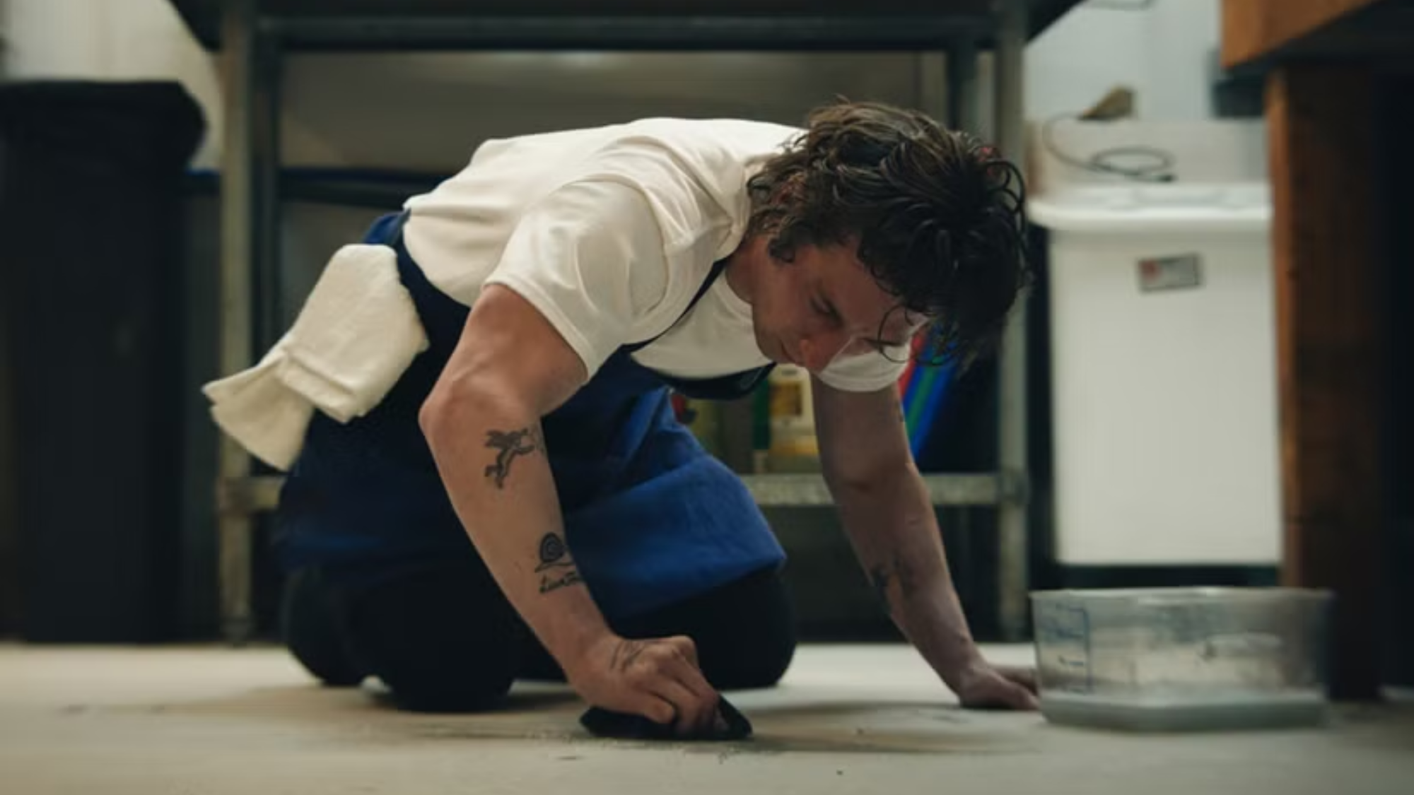 Carmy in a blue apron scrubbing the kitchen floor on his knees