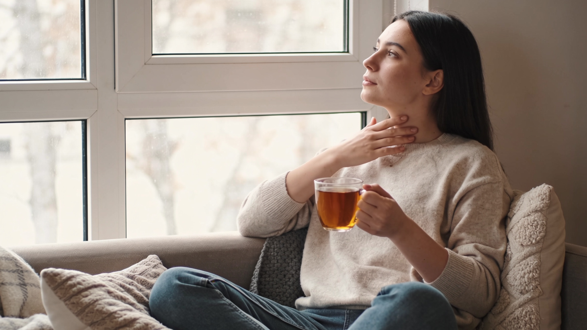 young woman sitting by a window with a cup of tea while gently touching her throat