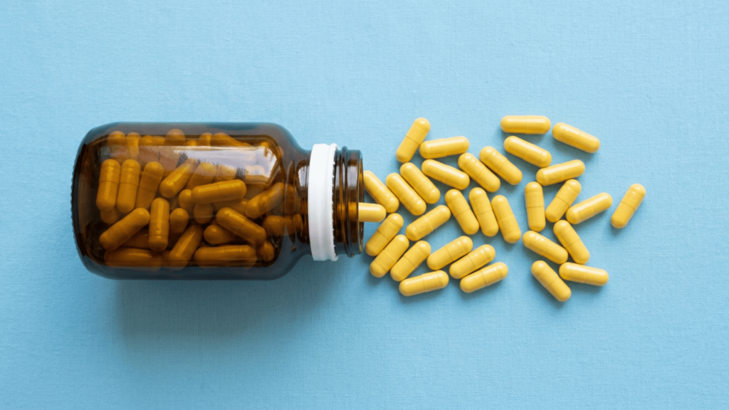 An amber glass supplement bottle on its side with yellow capsules spilling out on a light blue background