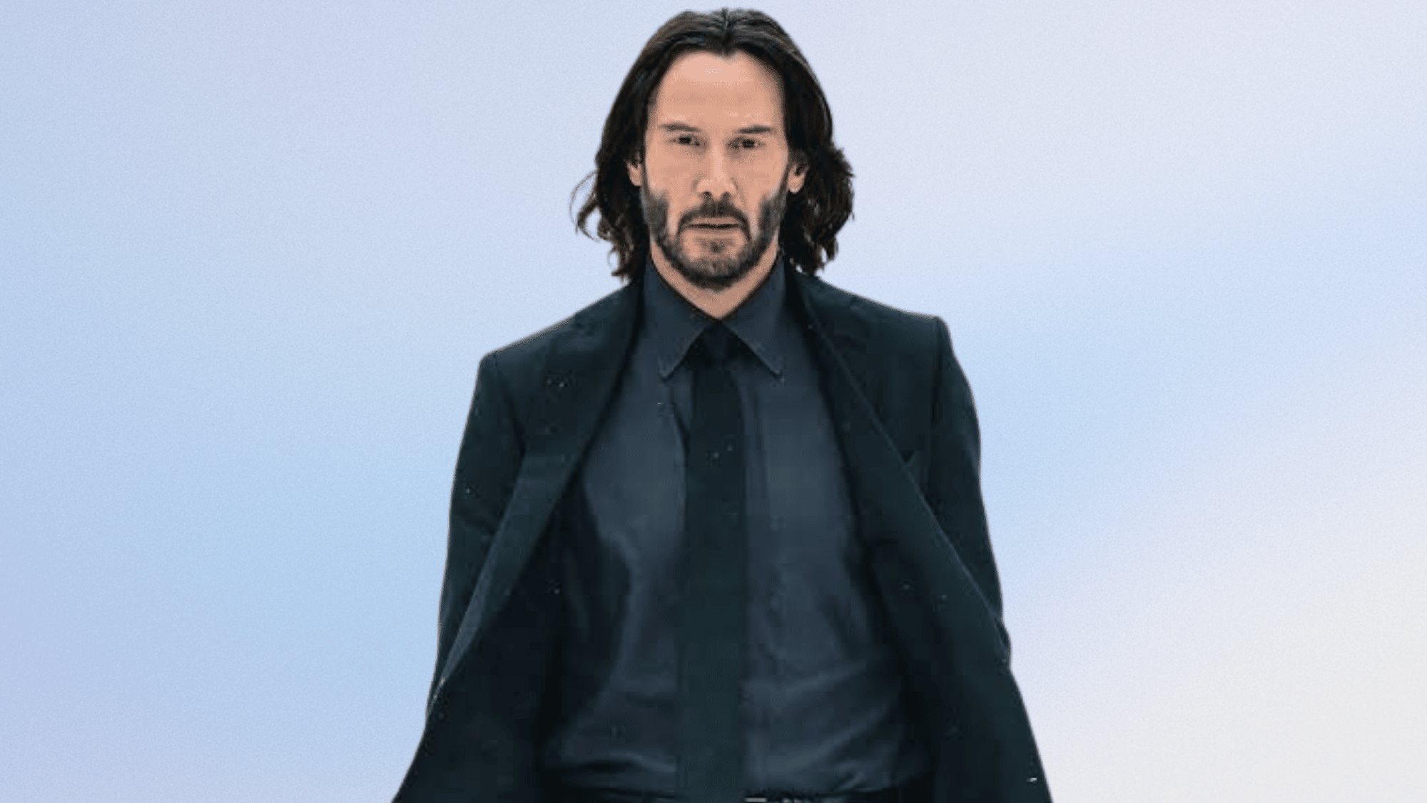 Actor in a black suit walking against a pale blue background.