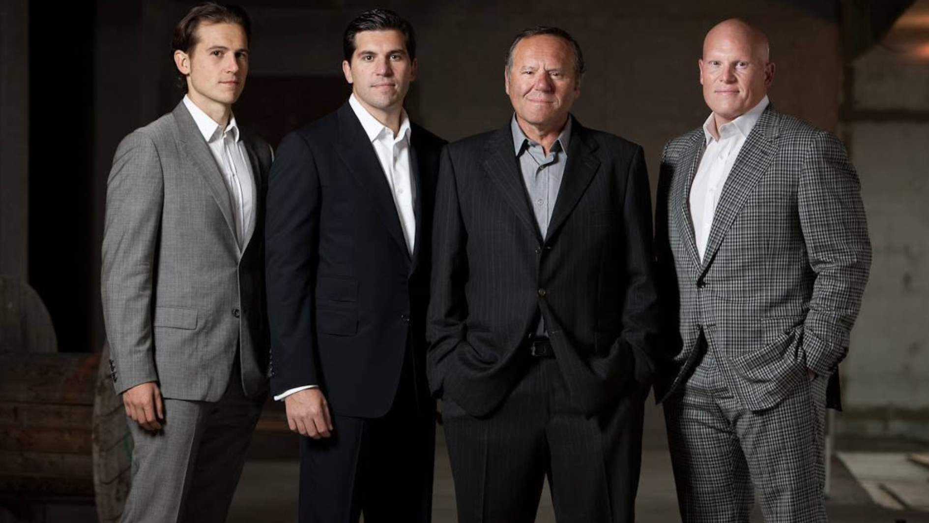 four men in formal suits pose together indoors against a dark, softly lit background