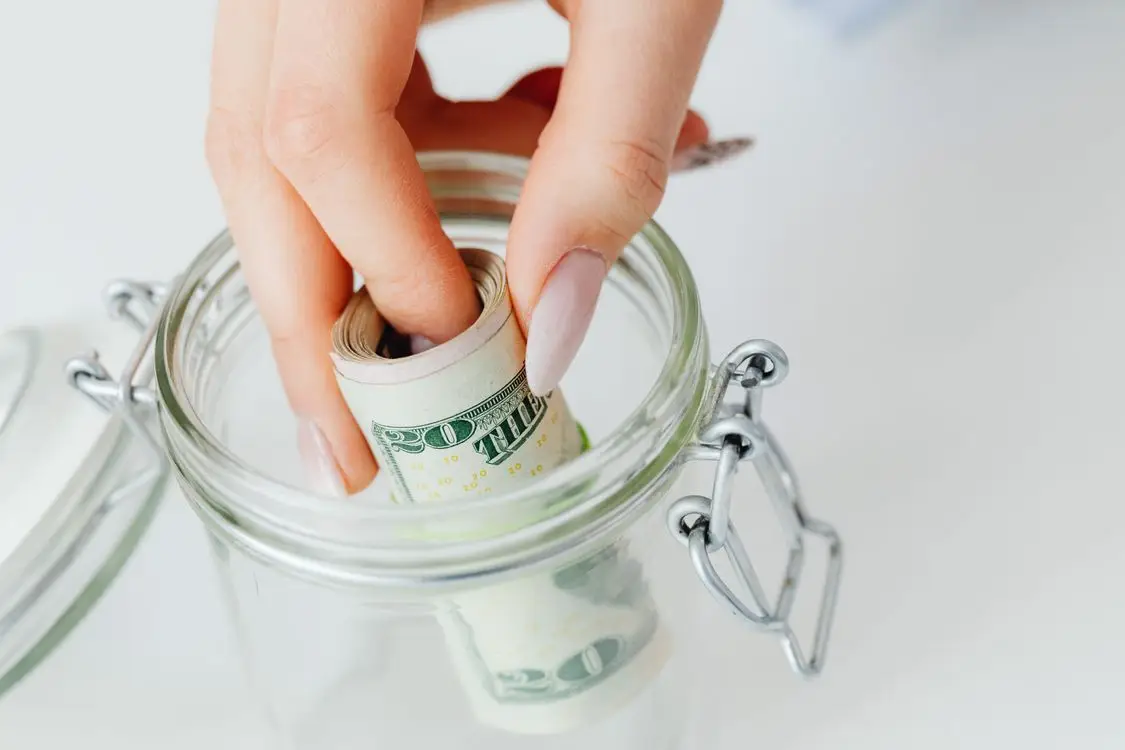 Hand placing rolled dollar bills into glass jar on white surface