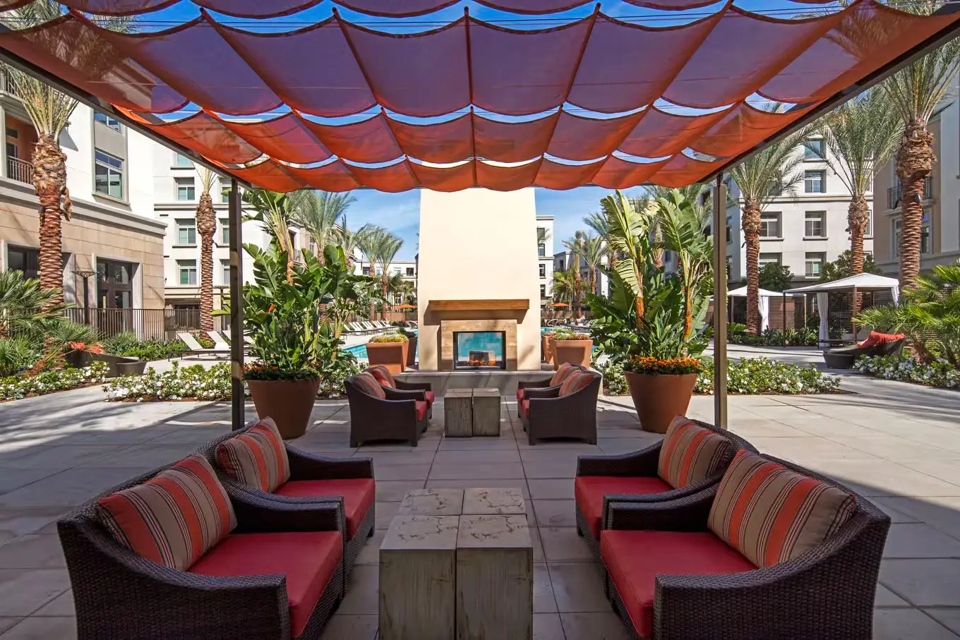 Outdoor lounge area with wicker furniture under red canopy, facing modern fireplace and lush plants