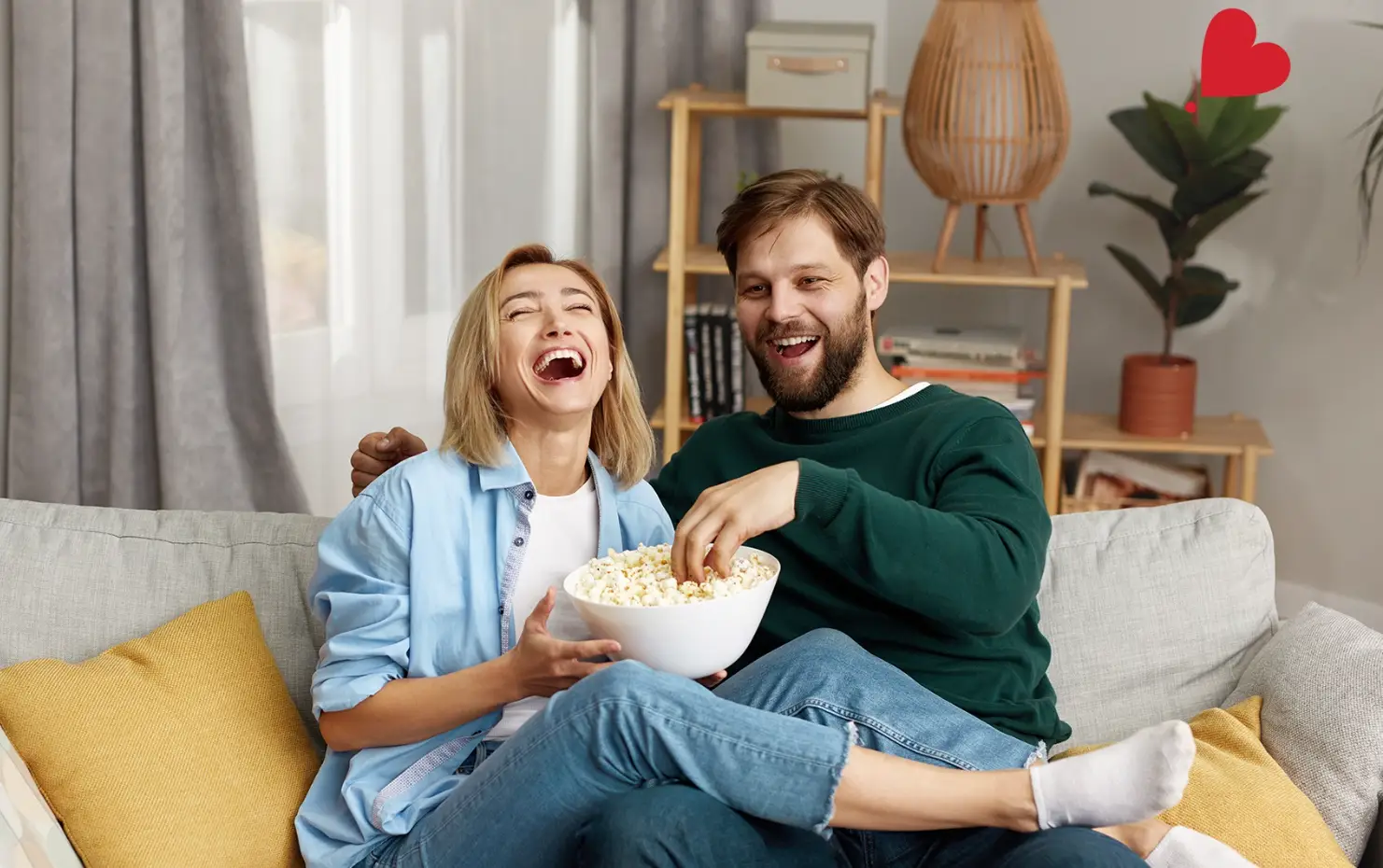 Couple laughing and sharing popcorn on a cozy couch with pillows in a living room