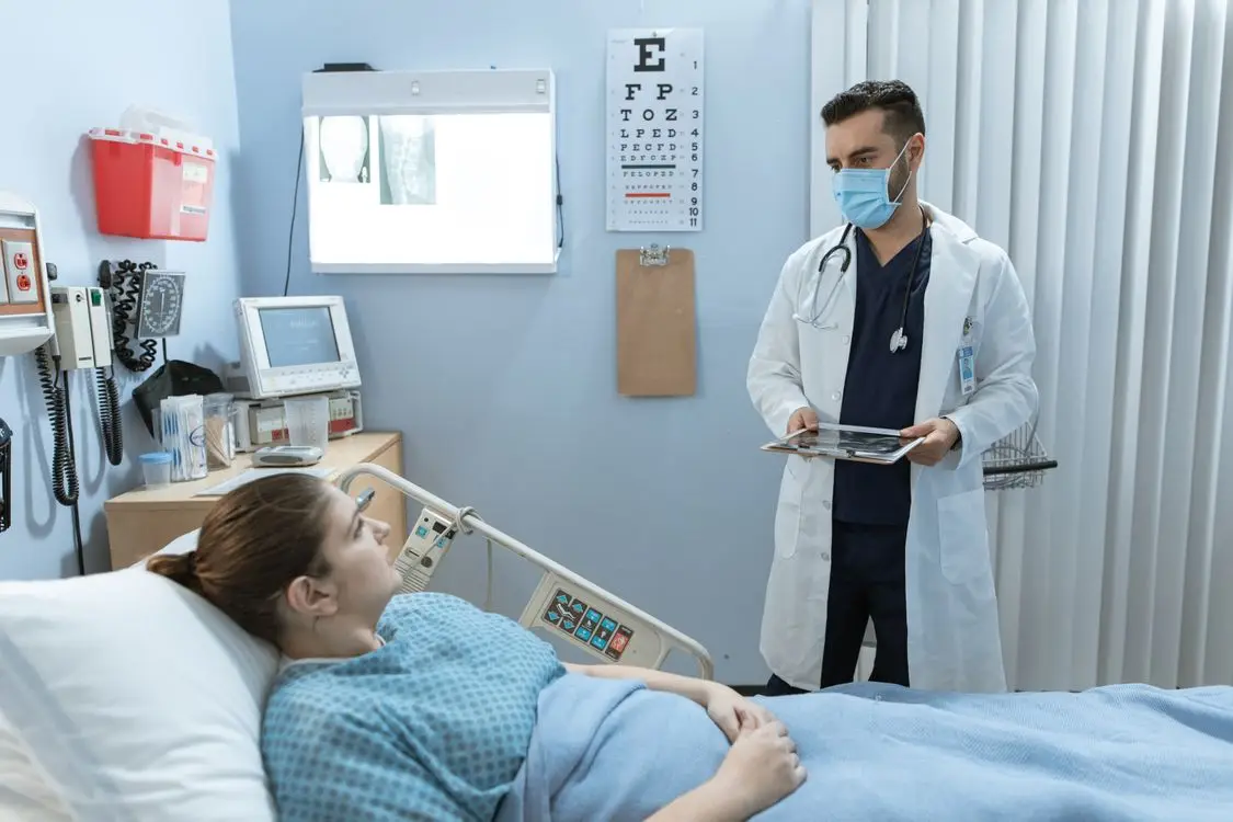 Doctor wearing a mask and holding a clipboard in a hospital room talking to a patient