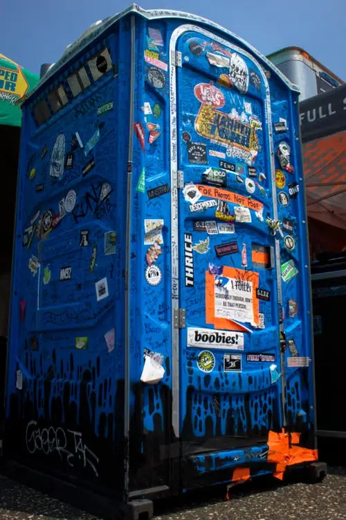 Blue portable toilet covered in colorful stickers and graffiti under bright sunlight