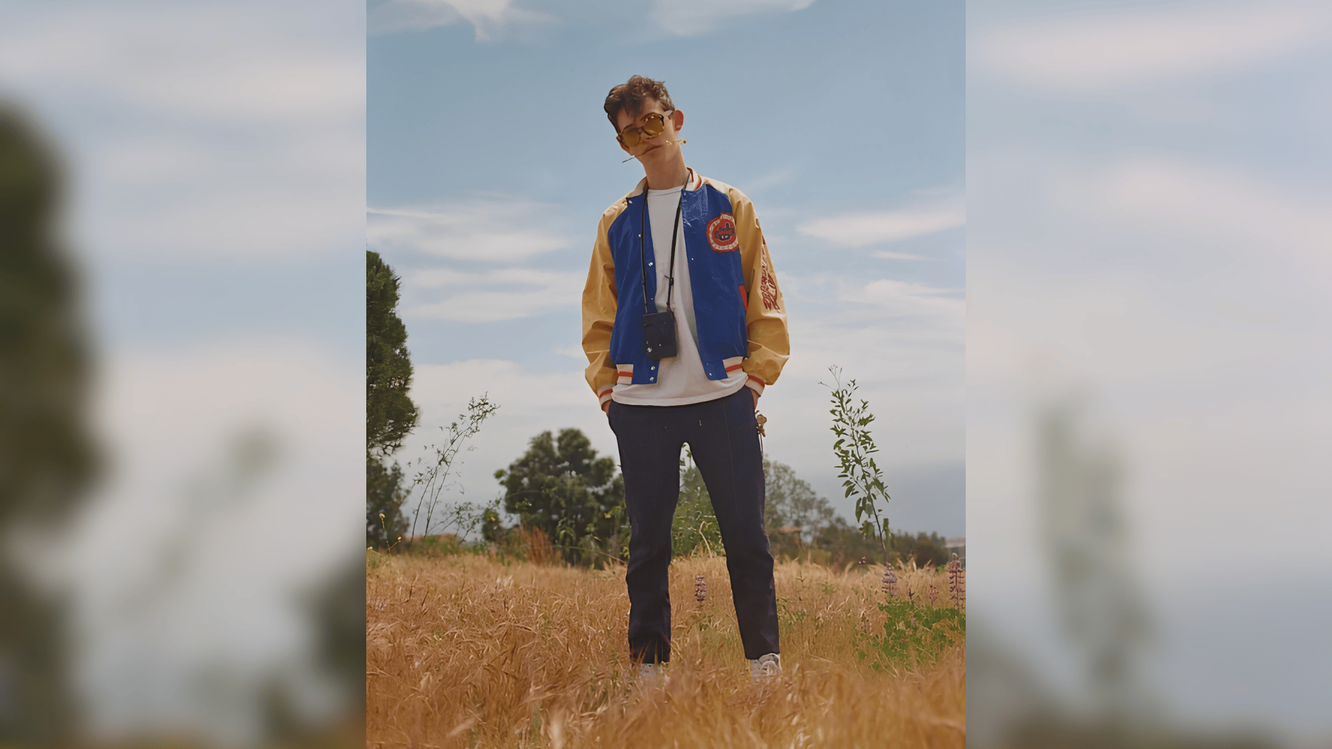 griffin musk standing in a field wearing a blue and gold varsity jacket and dark trousersYou said: alt text