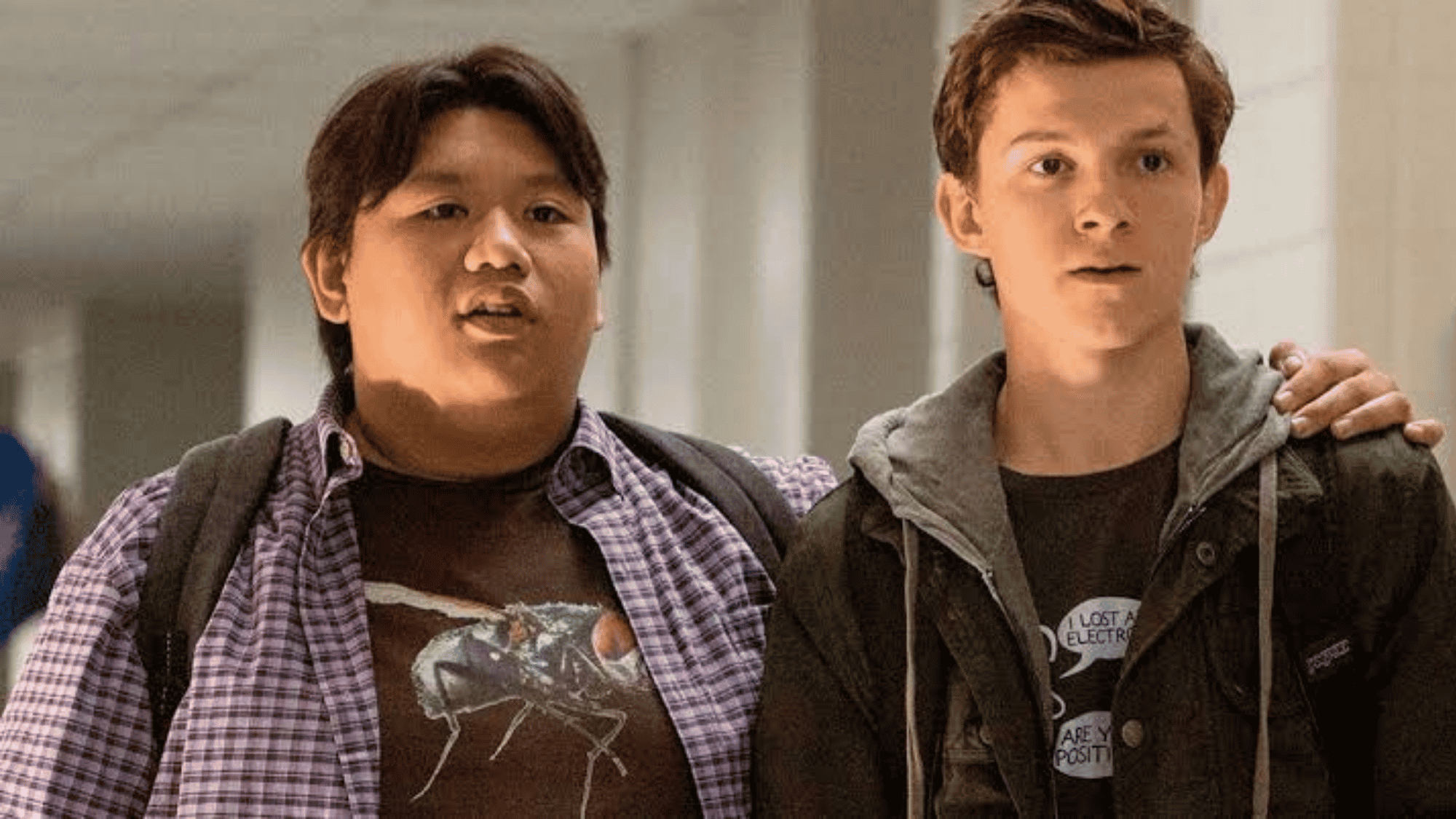 Two young actors in a school scene, one with dark hair standing beside his friend