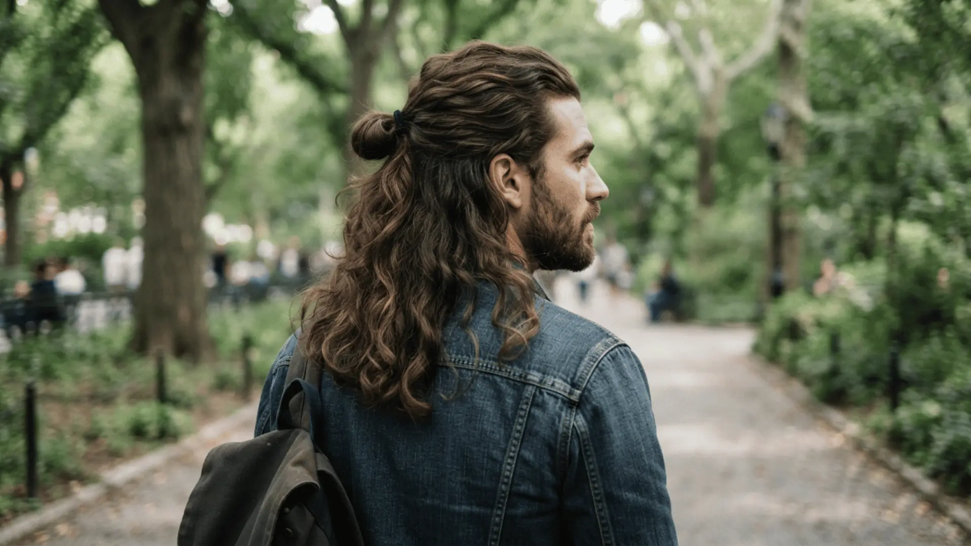man with long curly hair tied back walks through a leafy park wearing a denim jacket and carrying a bag
